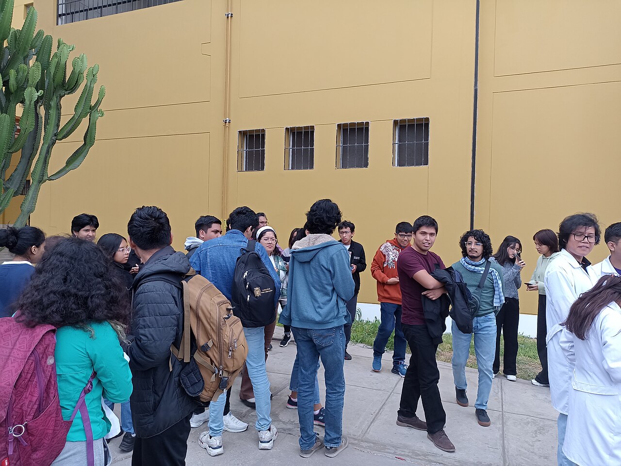 Students with backpacks and white lab coats waiting in a group beside a yellow university building in Lima during an evacuation drill