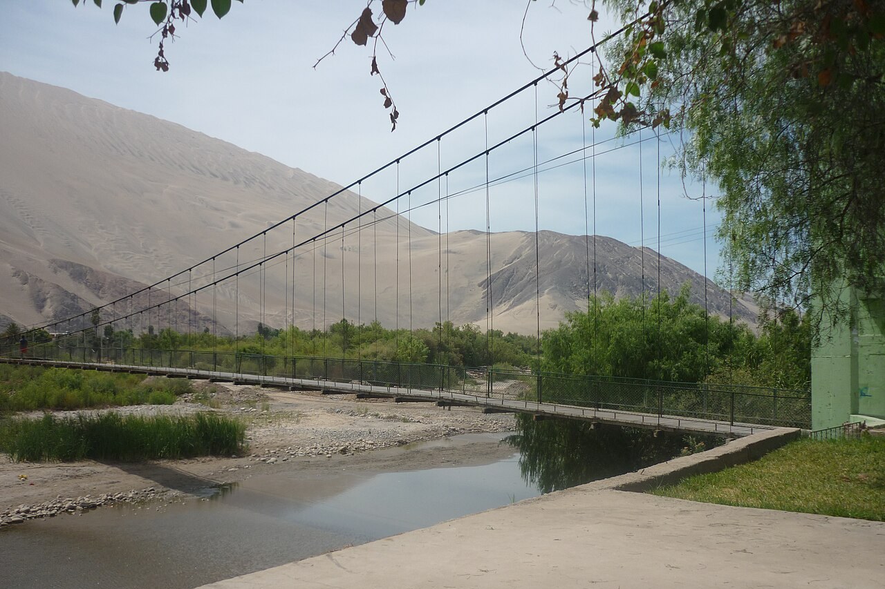 A pedestrian suspension bridge over the Acarí river below bare desert hills in Acarí, Arequipa, Peru