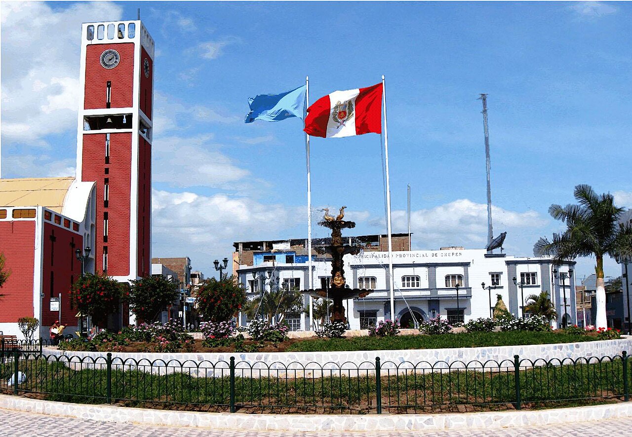 A red clock tower, a fountain and the white municipal building of Chepén behind Peruvian and regional flags under a blue sky