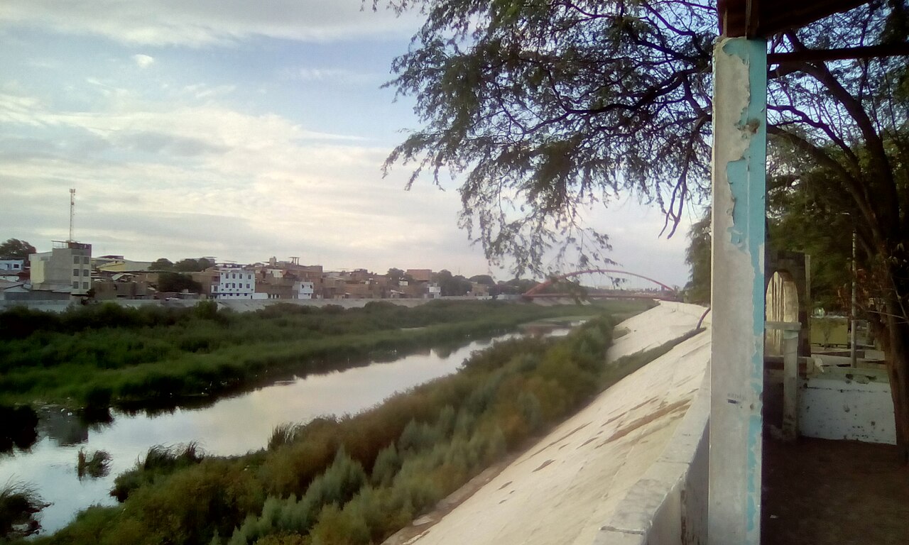 The Piura River at low water between concrete flood walls, with green banks, houses and a red footbridge in the distance