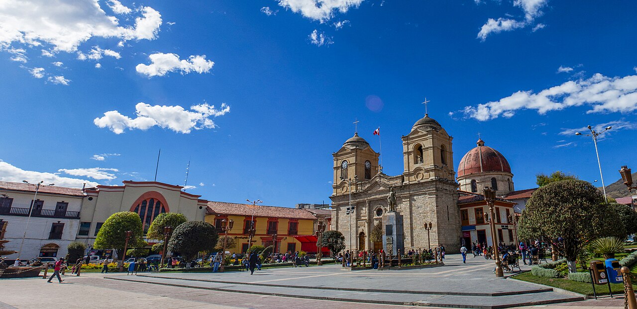 The twin-towered stone cathedral of Huancayo beside a red-domed church on a wide square with gardens, lamp posts and people walking under a blue sky.
