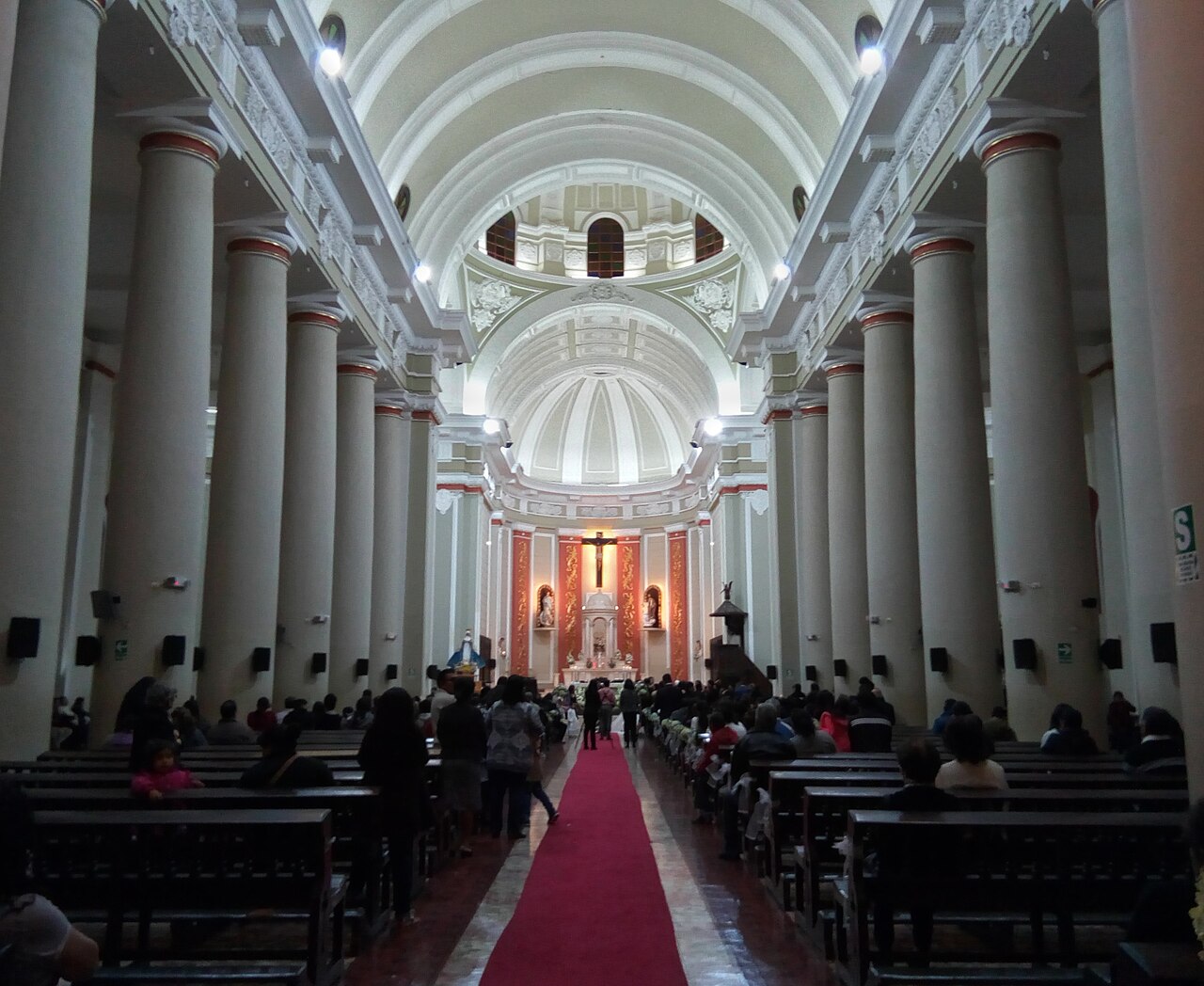 The long white nave of a Peruvian cathedral with columns, a red carpet and worshippers on wooden pews facing the altar