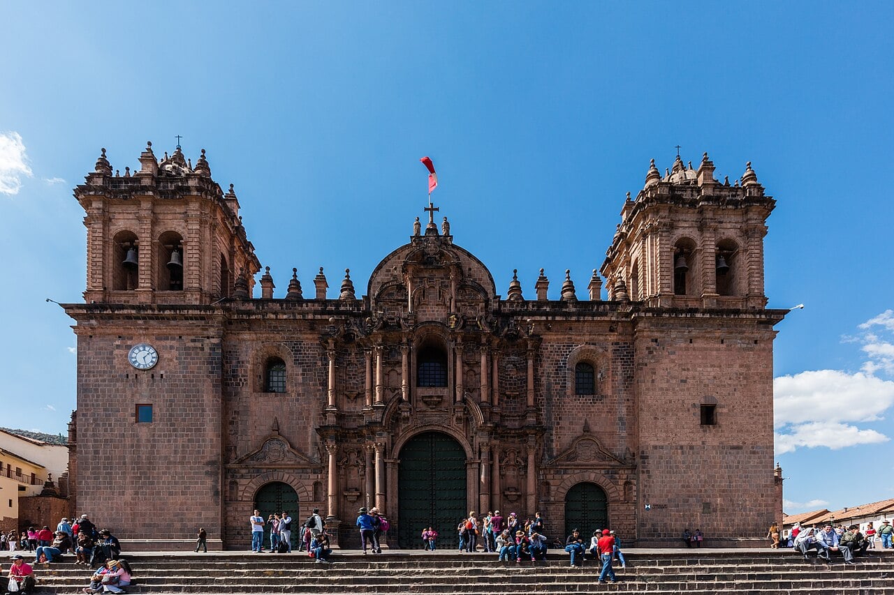 Stone facade and twin bell towers of Cusco Cathedral on the Plaza de Armas, with people sitting on the steps under a blue sky