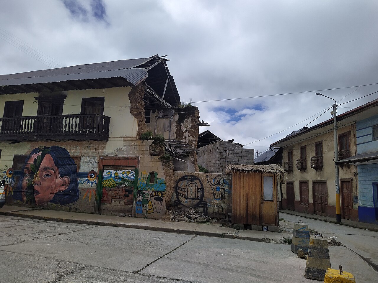 Two-storey house with a wooden balcony and a partly collapsed wall, its ground floor covered in painted murals, on a street corner in Cerro de Pasco