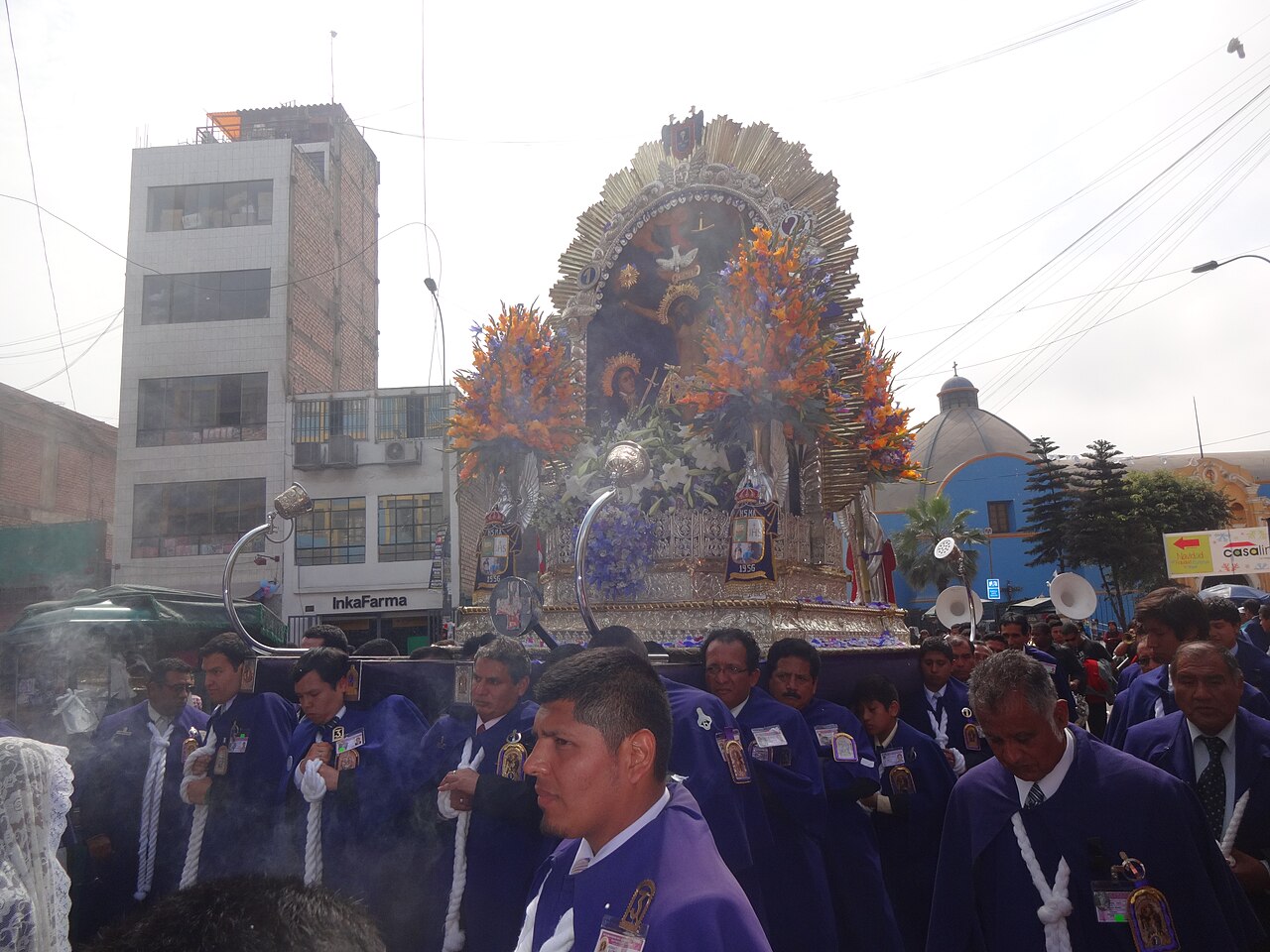 Men in purple robes with white cords carry a silver-framed image of Christ, decorated with orange and purple flowers, through a street in Lima.