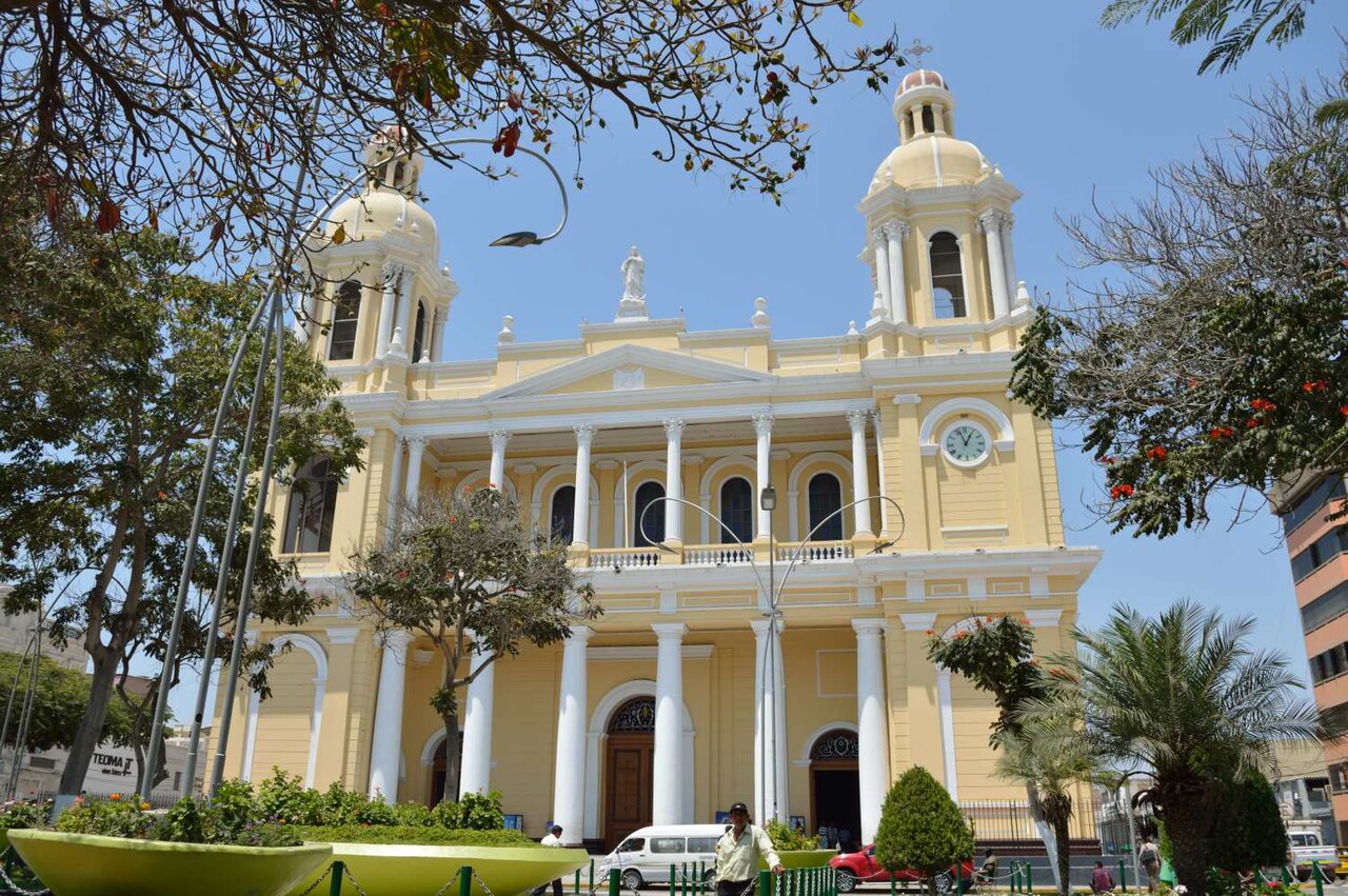 The yellow and white neoclassical facade of Chiclayo Cathedral, with two bell towers, a clock and white columns, seen across a square with trees.