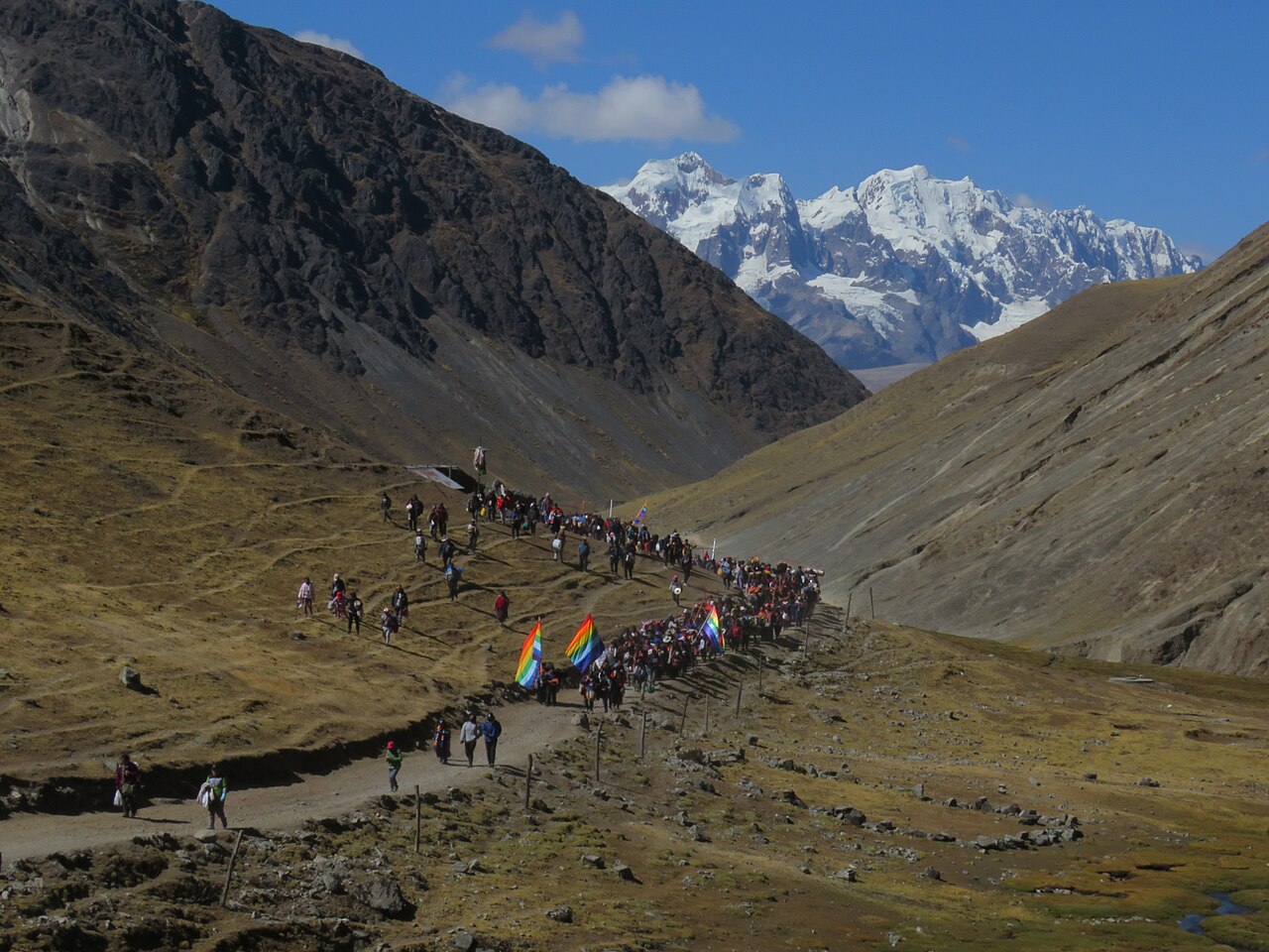 A long line of pilgrims carrying rainbow-striped flags walks up a dry Andean valley towards snow-capped mountains under a blue sky.