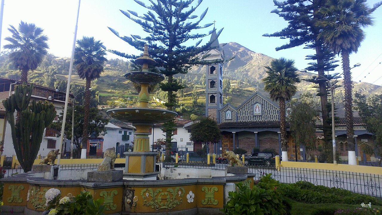 Main square of San Marcos with an ornate fountain, palm and araucaria trees, and the parish church tower against a mountain