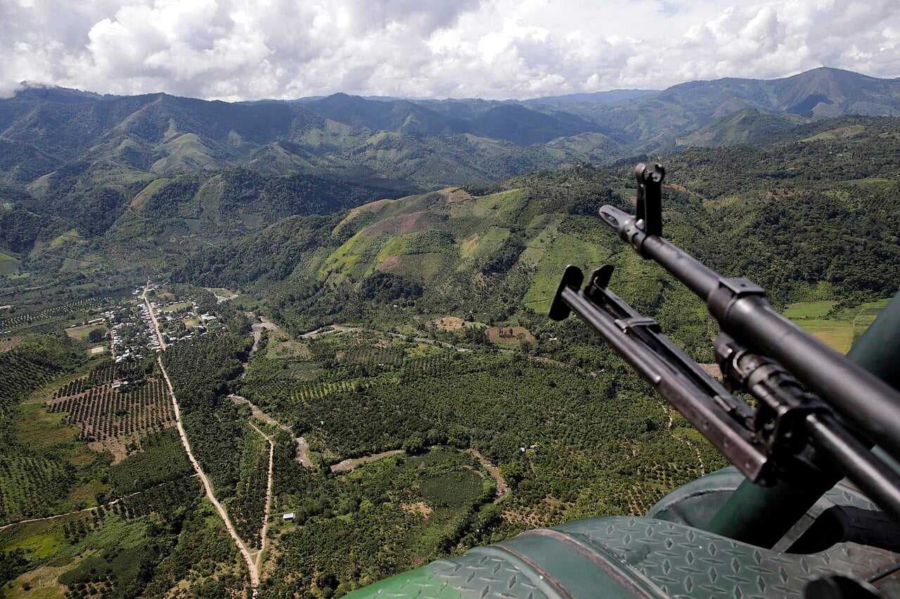 Aerial view of green valleys, farm plots and a small town in Peru, with a mounted machine gun on a helicopter in the foreground