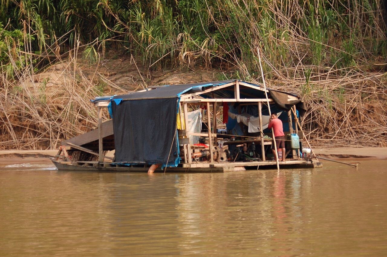 A floating gold-mining dredge raft with a worker under a tarpaulin roof on the Tambopata River, Madre de Dios, Peru