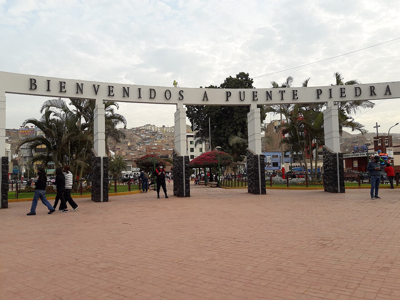 White arch reading Bienvenidos a Puente Piedra over a brick plaza with palm trees, pedestrians and a police officer, with hillside houses behind
