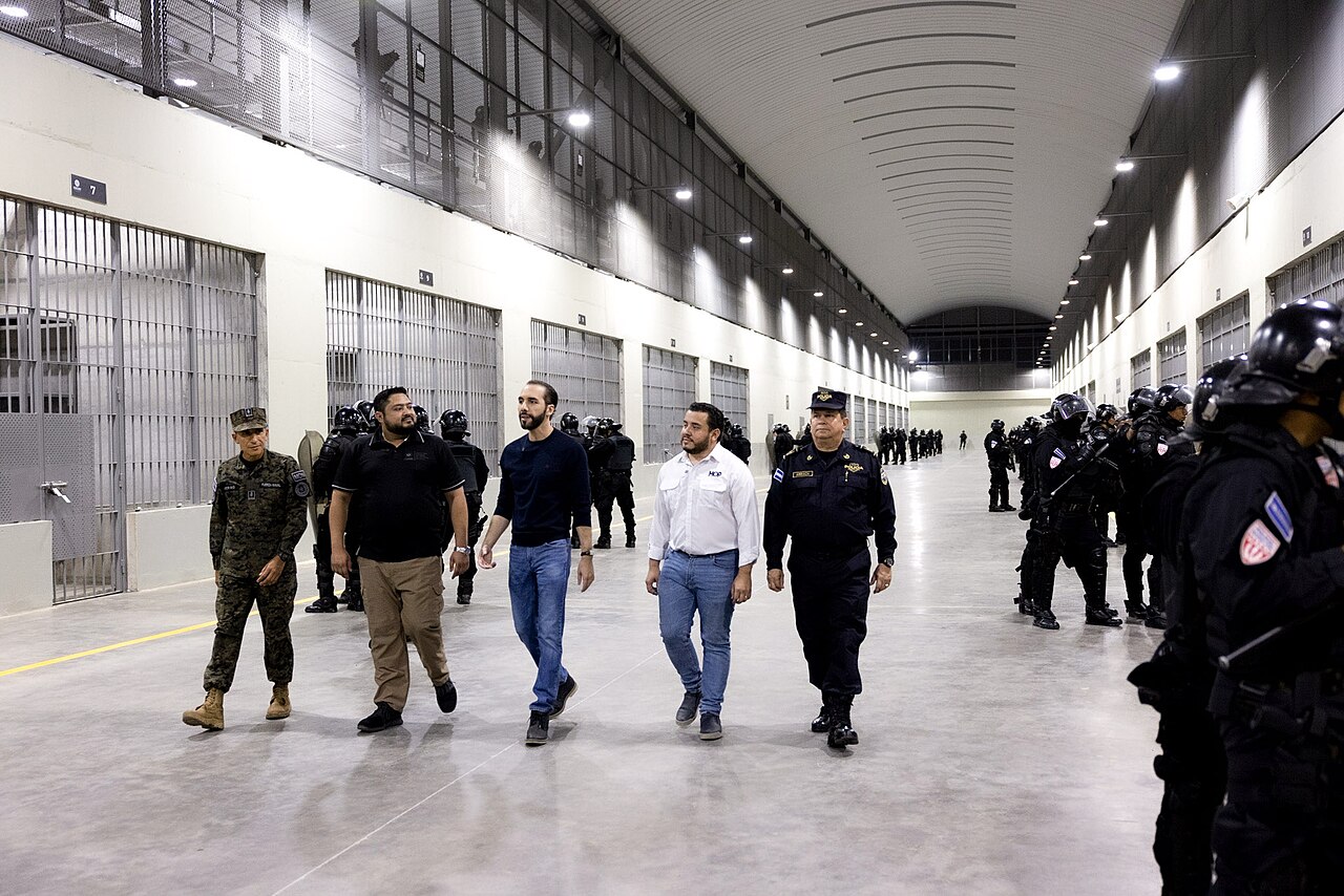 Five men walk along a long cell-block corridor at CECOT with barred cell doors on the left and rows of riot police on the right