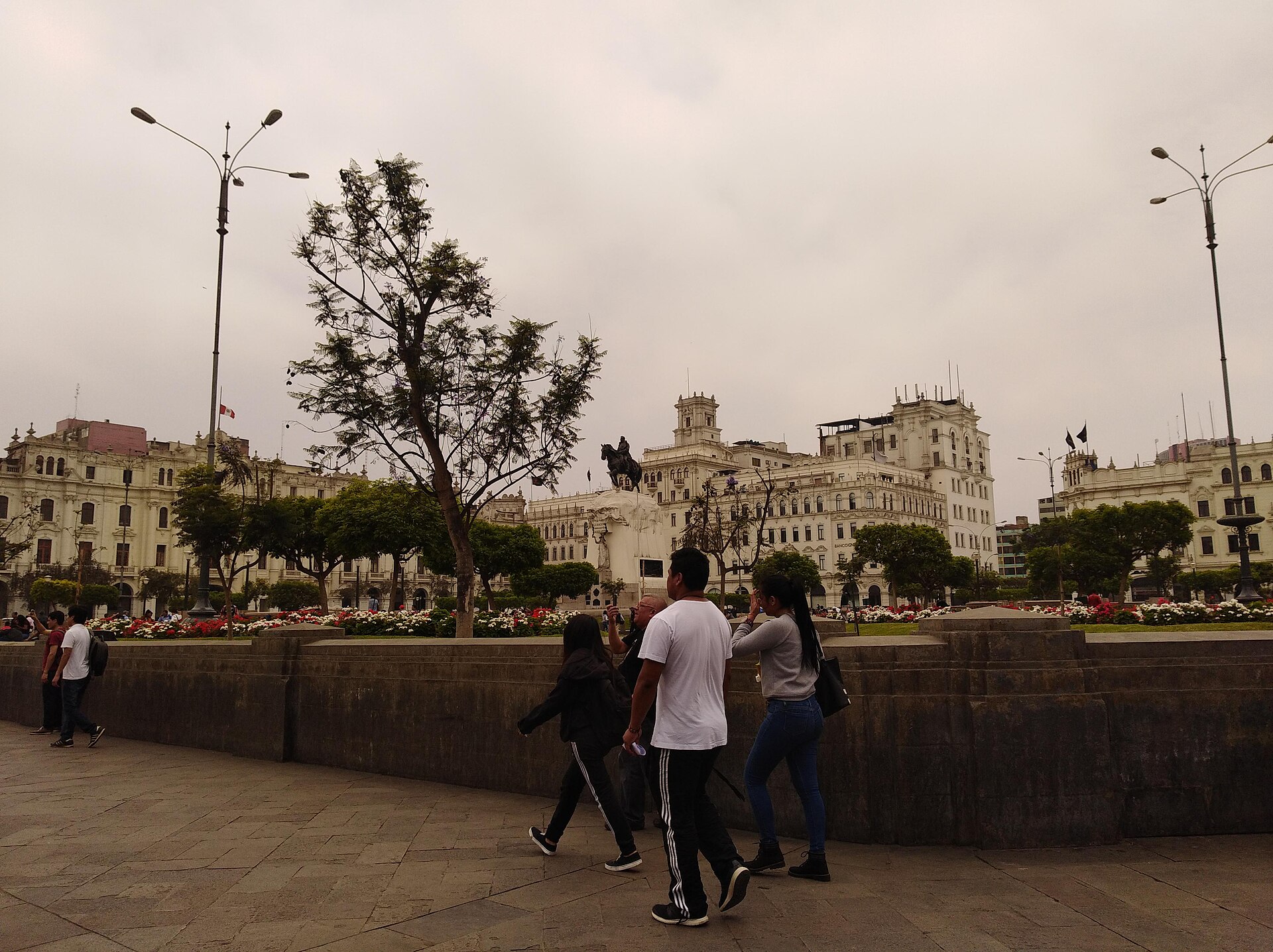 Pedestrians walk past Plaza San Martín under an overcast sky in central Lima