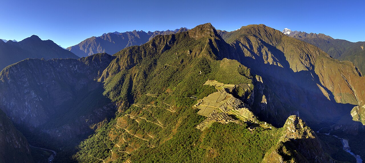 Aerial-style view of Machu Picchu's terraces on a green mountain ridge surrounded by Andean peaks and a river valley
