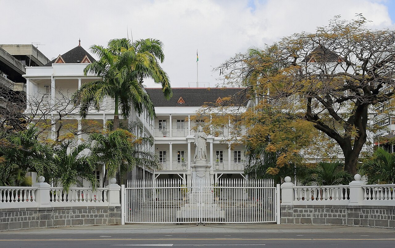 Government House, a white colonial-style building behind a gate and palm trees in Port Louis, Mauritius.