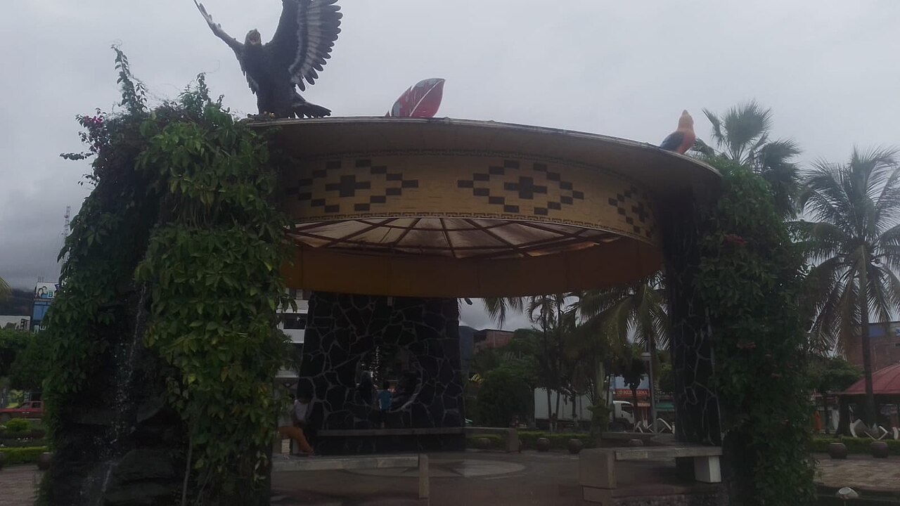 A round pavilion with a large bird sculpture and a patterned roof, framed by vines and palm trees, in a town square