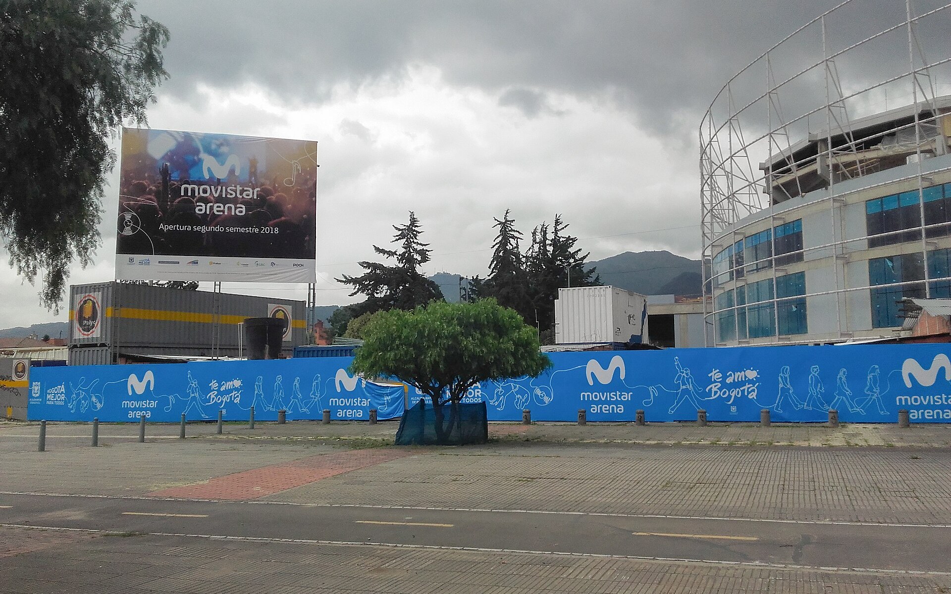 Movistar Arena in Bogotá, Colombia, under construction in April 2018, behind blue hoardings and a billboard announcing its opening