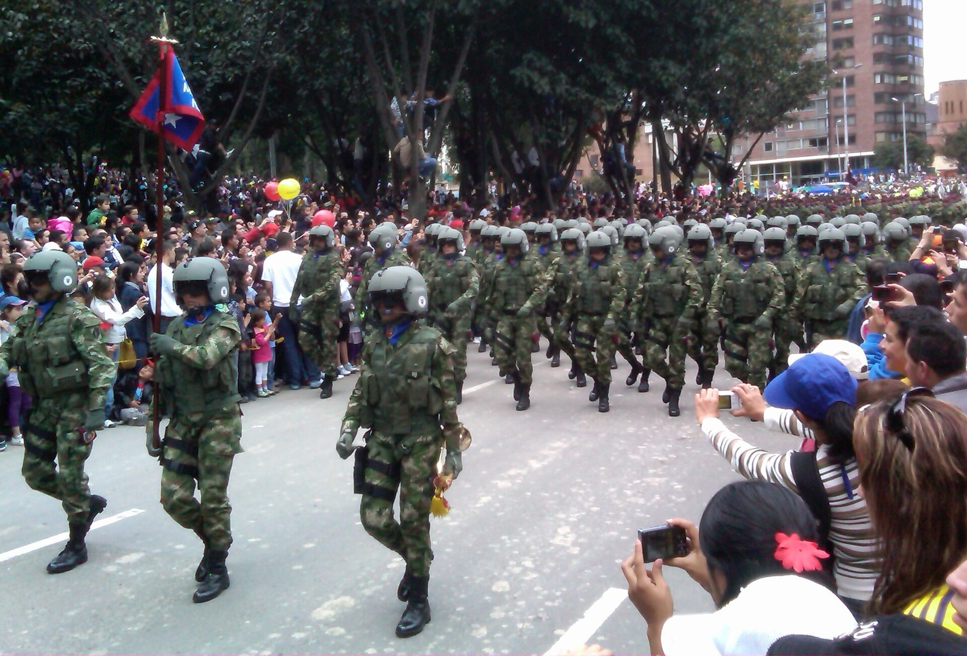 Colombian army aviation troops in camouflage and helmets march in formation past crowds of spectators taking photos, led by a flag bearer