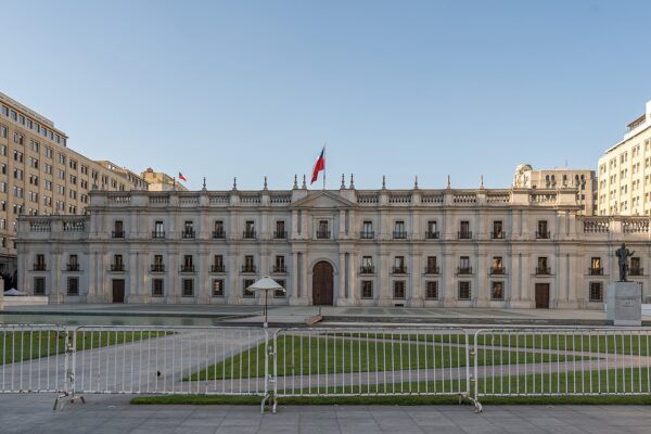The white neoclassical facade of La Moneda Palace in Santiago with a Chilean flag on the roof, a lawn and metal barriers in front