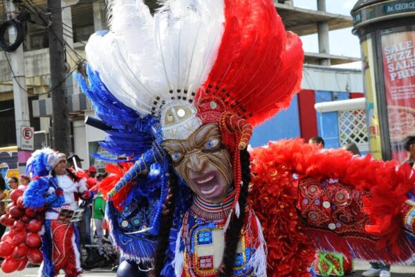 A carnival performer in a red, white and blue feathered diablo cojuelo costume and a devil mask with gold markings on a city street, with other costumed performers behind