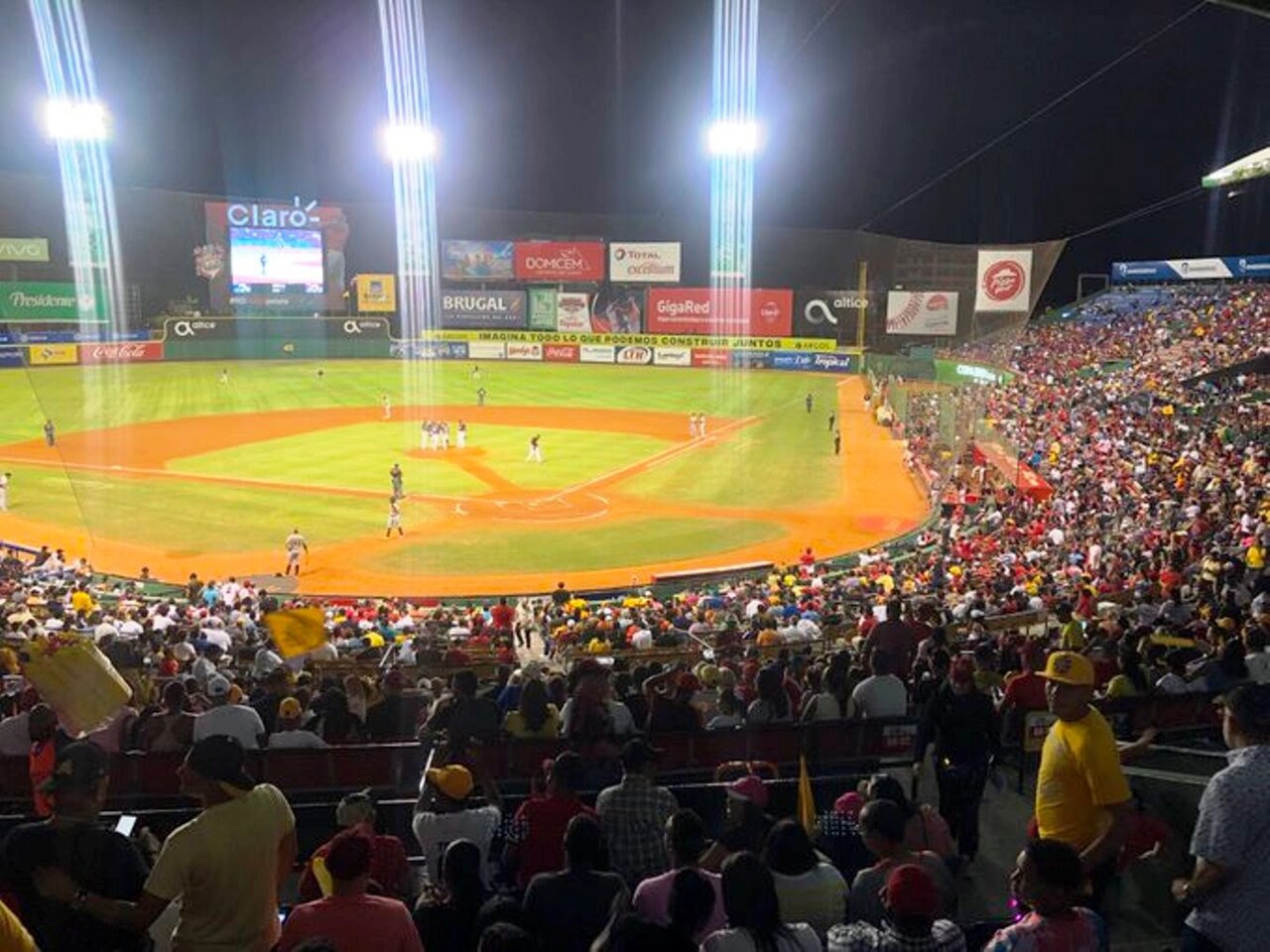 A floodlit baseball diamond seen from crowded stands at night, with sponsor boards and a scoreboard beyond the outfield