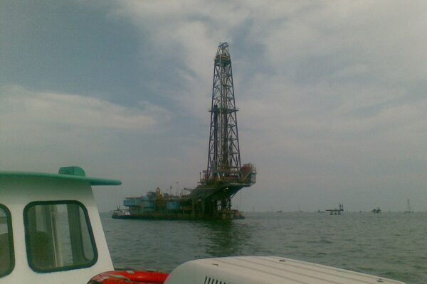 A tall offshore drilling rig on a platform in the grey-green water of Lake Maracaibo, seen from the bow of a small boat under a cloudy sky
