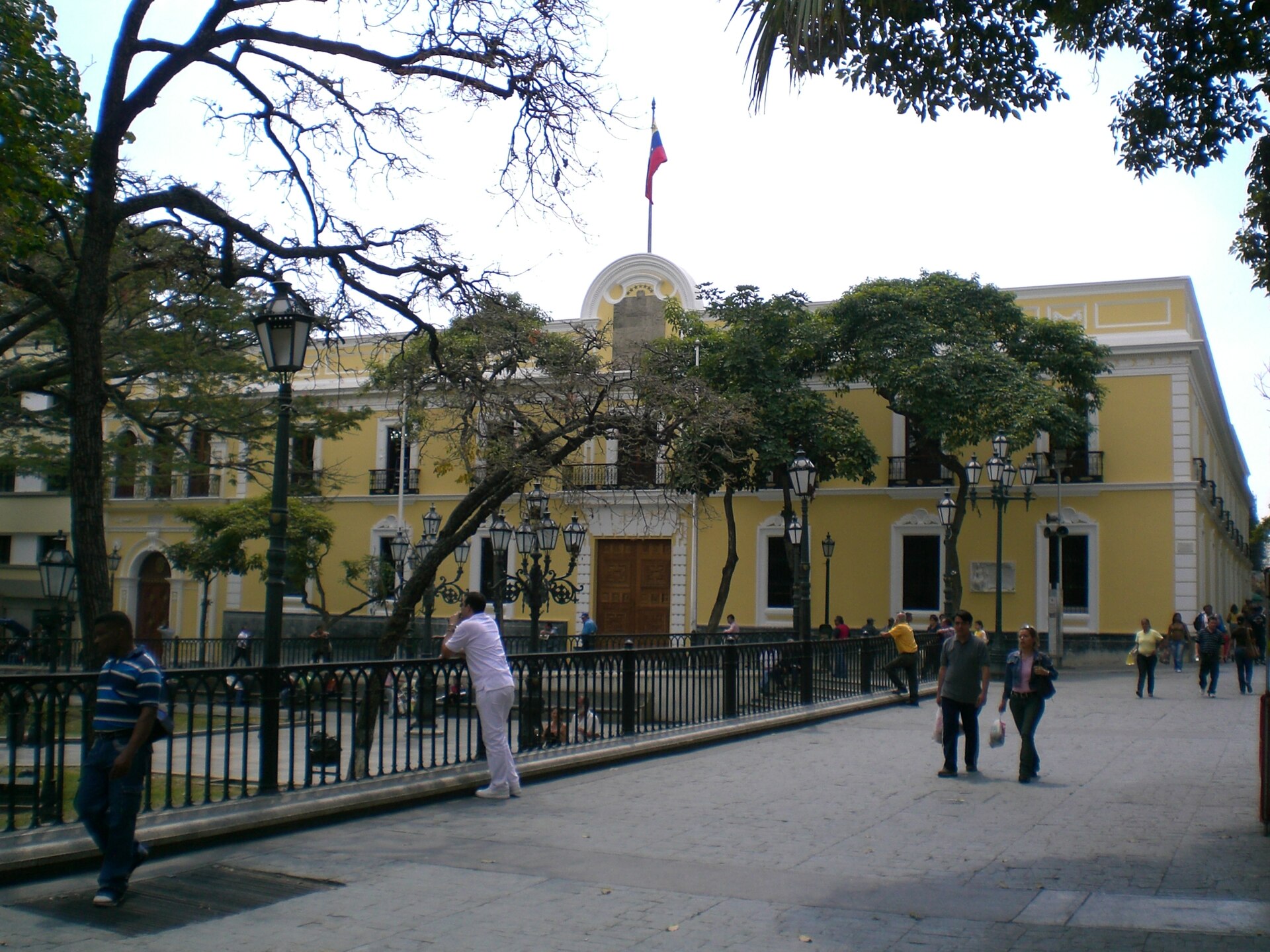 The yellow colonial facade of the Casa Amarilla in Caracas, with a Venezuelan flag on the roof, trees and people walking on the square in front