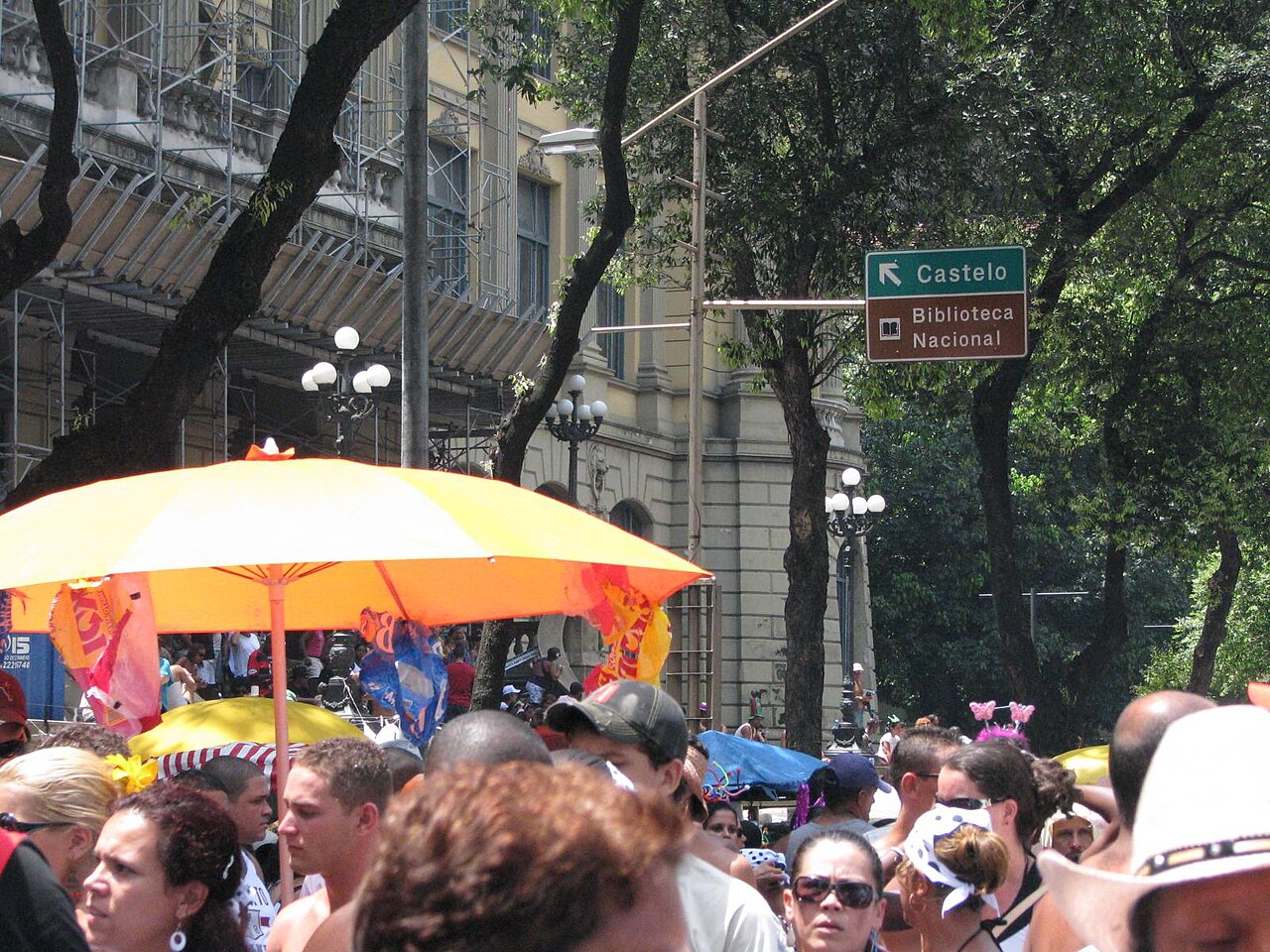 Crowd of revellers under an orange umbrella in a tree-lined street beside a sign for the Biblioteca Nacional in central Rio