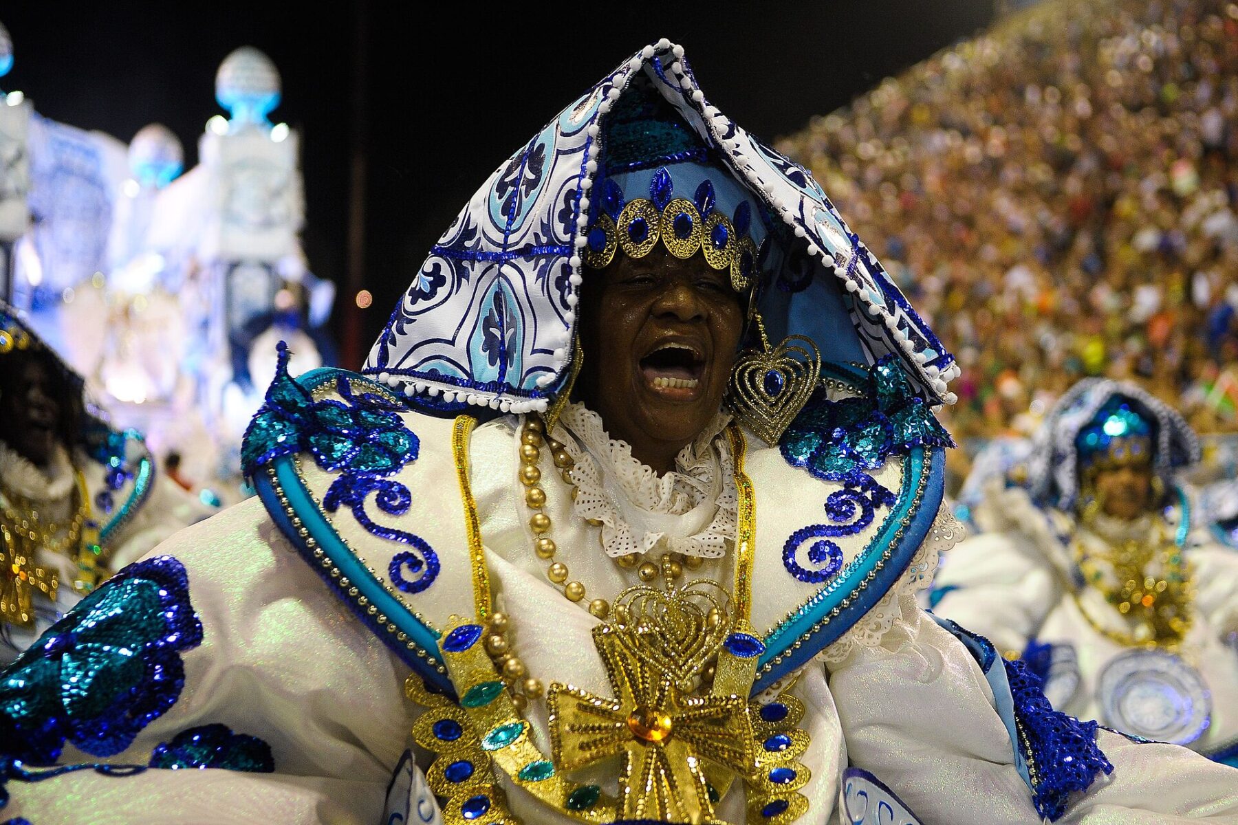 A samba school dancer in a white, blue and gold Baiana costume sings during a night parade, with packed stands behind