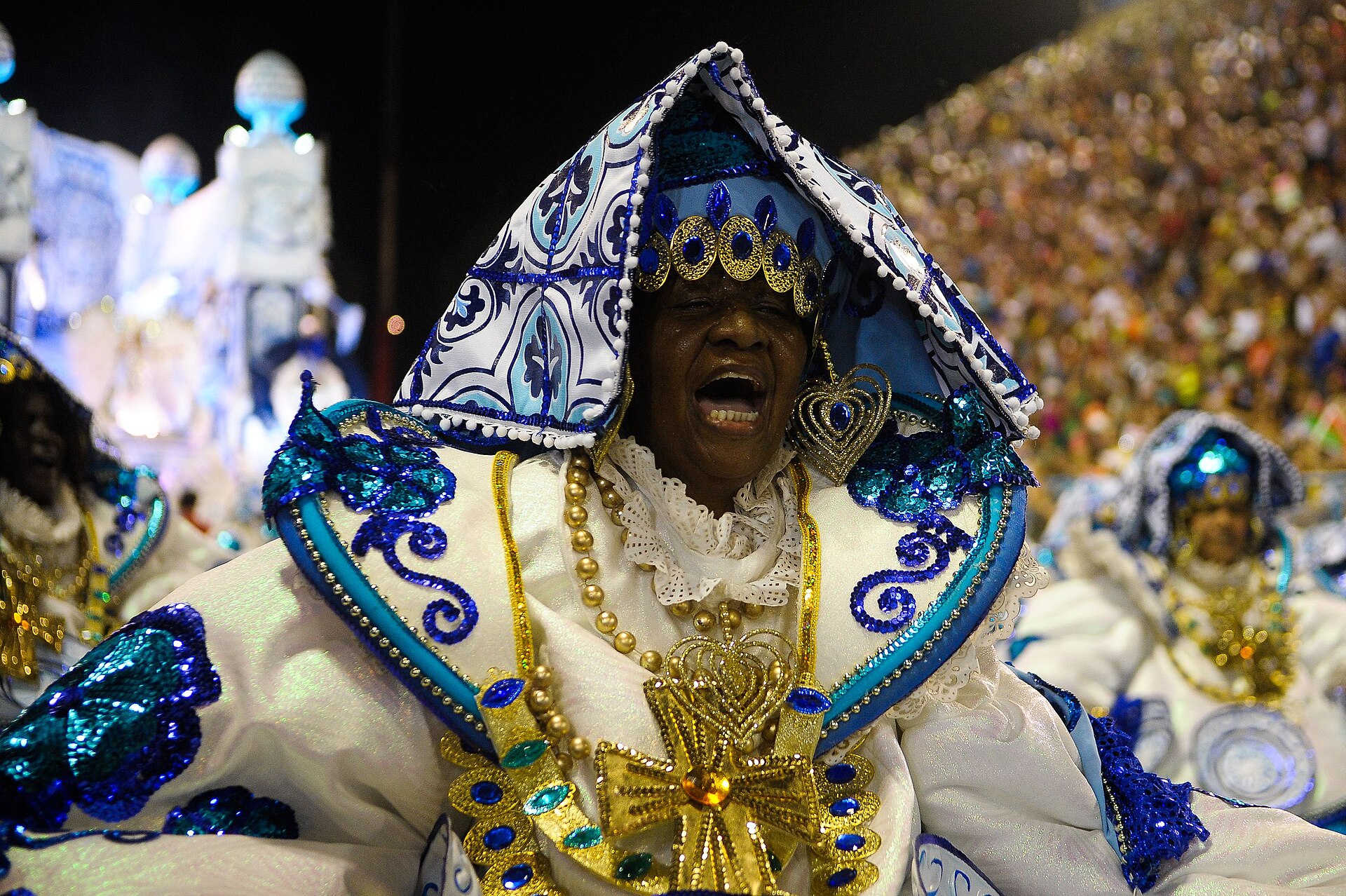 A samba school dancer in a white, blue and gold Baiana costume sings during a night parade, with packed stands behind