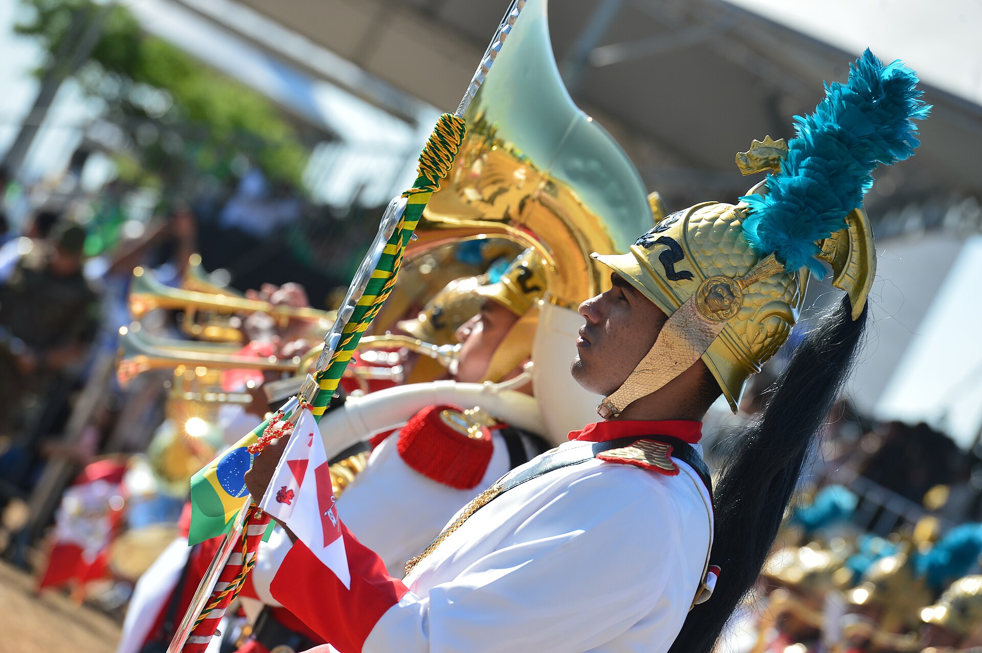 A young man in a gold helmet with a turquoise plume and a red and white uniform holds a lance with a Brazilian flag pennant beside brass musicians