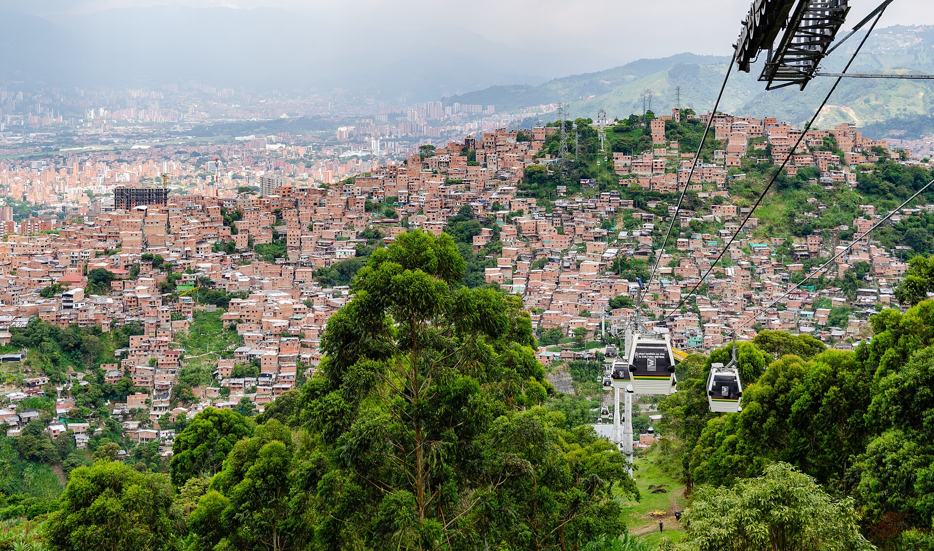 Cable car gondolas above green trees and a hillside packed with red-brick houses, with the Medellín valley in the background