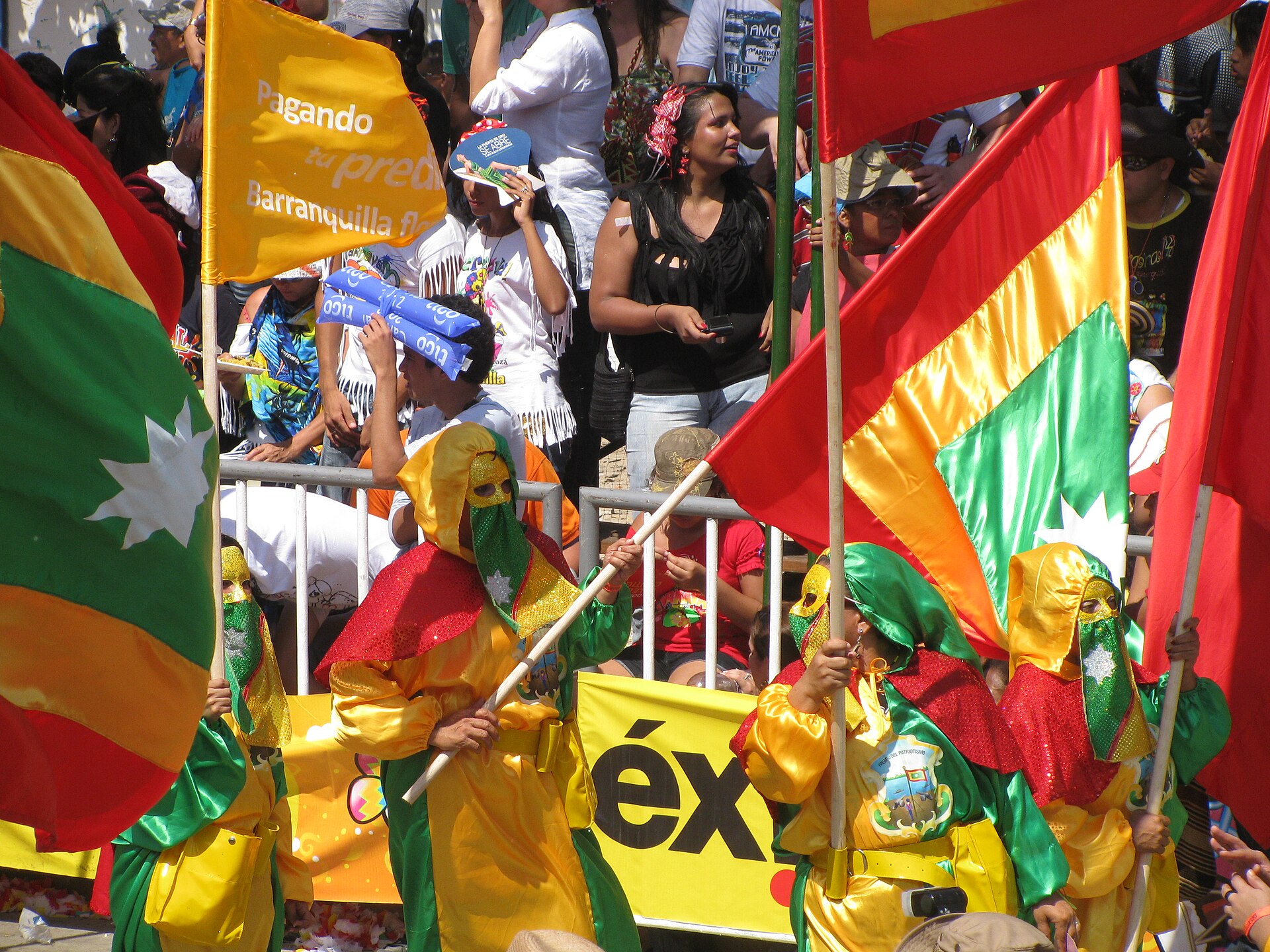 Masked performers in yellow, green and red costumes carrying large flags past a crowd of spectators behind a barrier