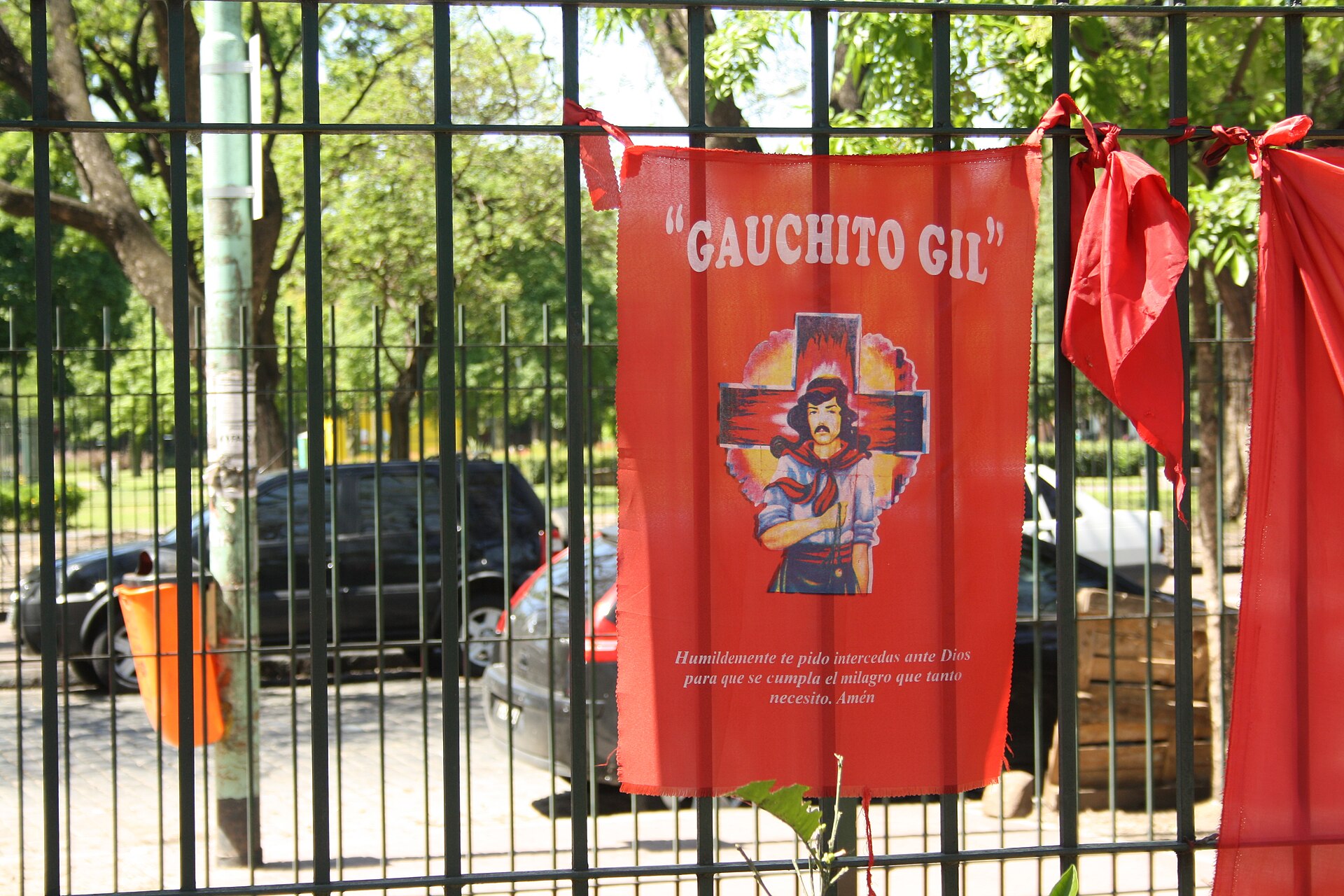 A red banner with the image of the Gauchito Gil and red ribbons tied to a metal fence in a park