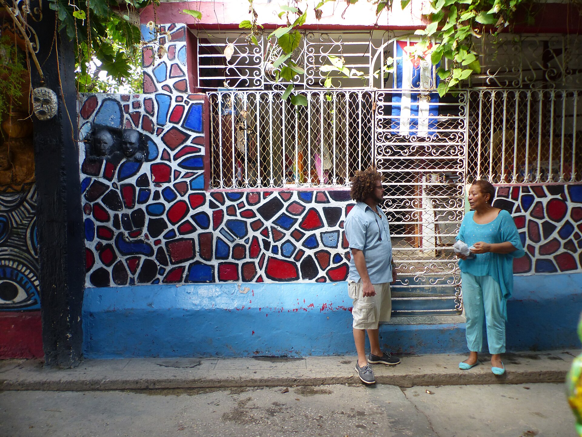 A man and a woman talking on the pavement in front of a house wall painted in red, blue and black mosaic patterns with a barred window