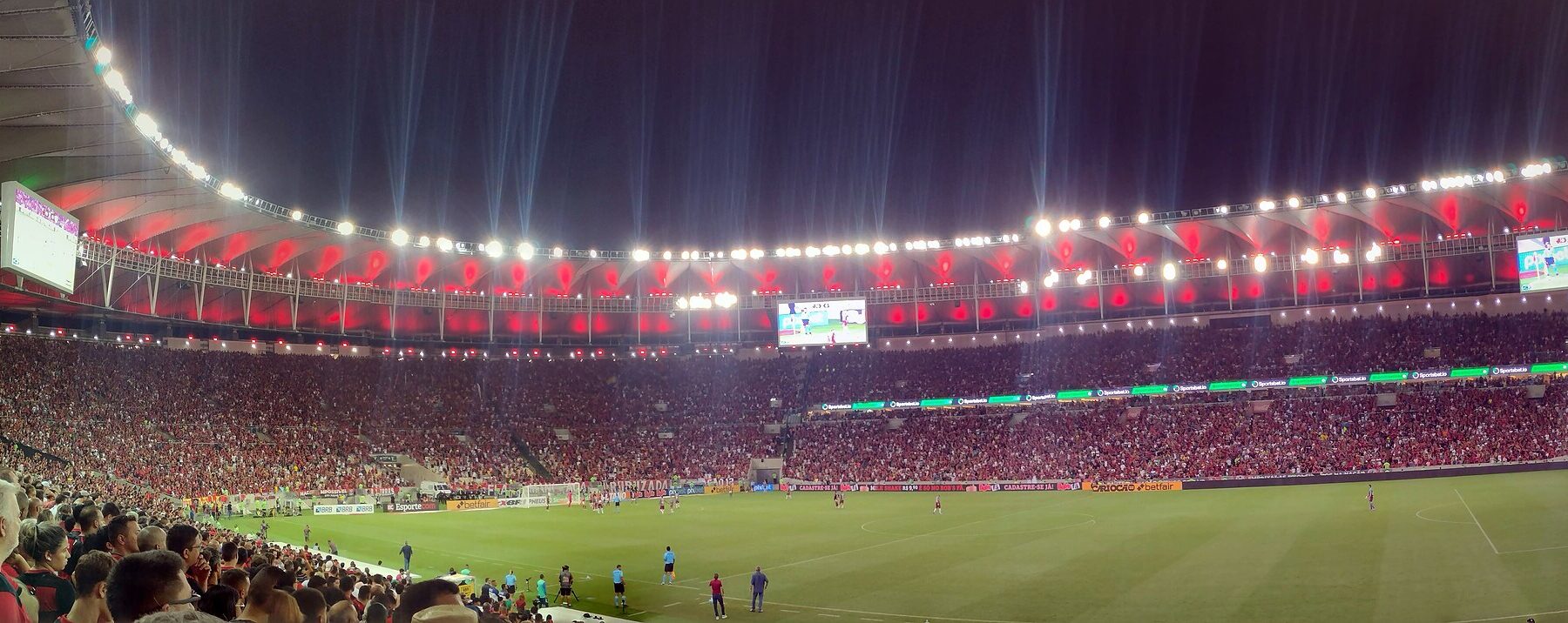 Floodlit Maracanã stadium packed with fans in red during a night match, seen from the lower stand