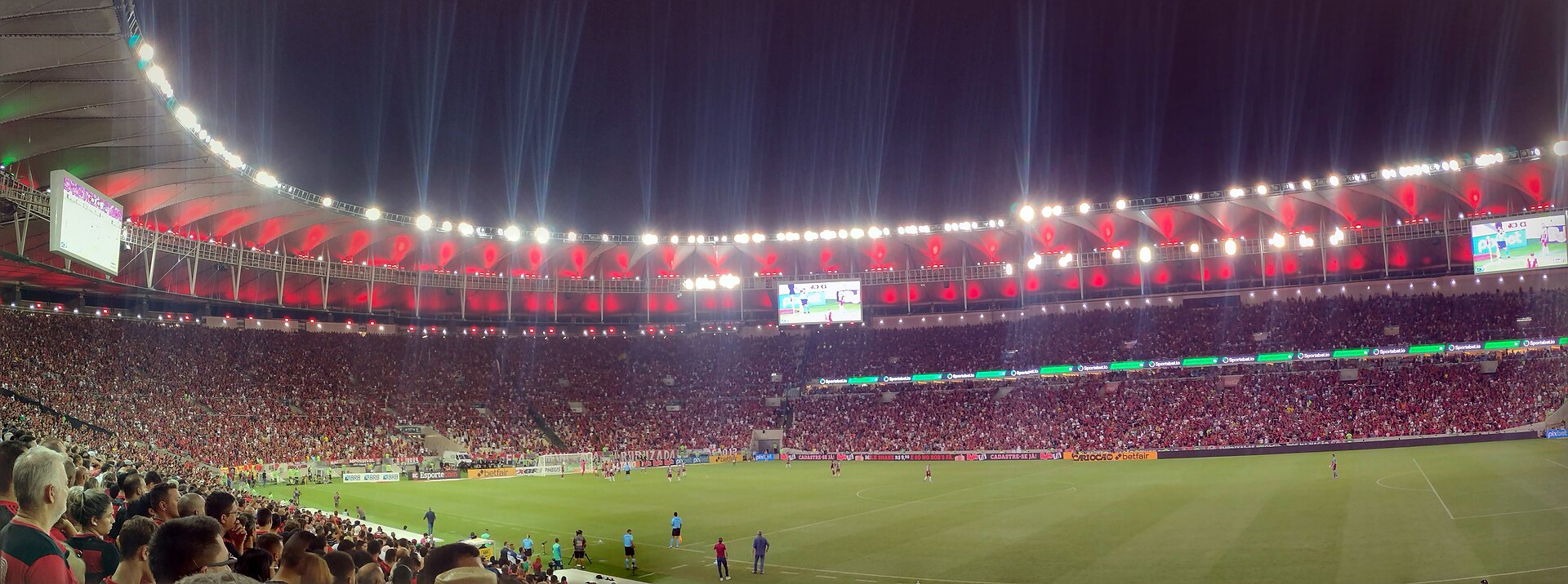 Floodlit Maracanã stadium packed with fans in red during a night match, seen from the lower stand