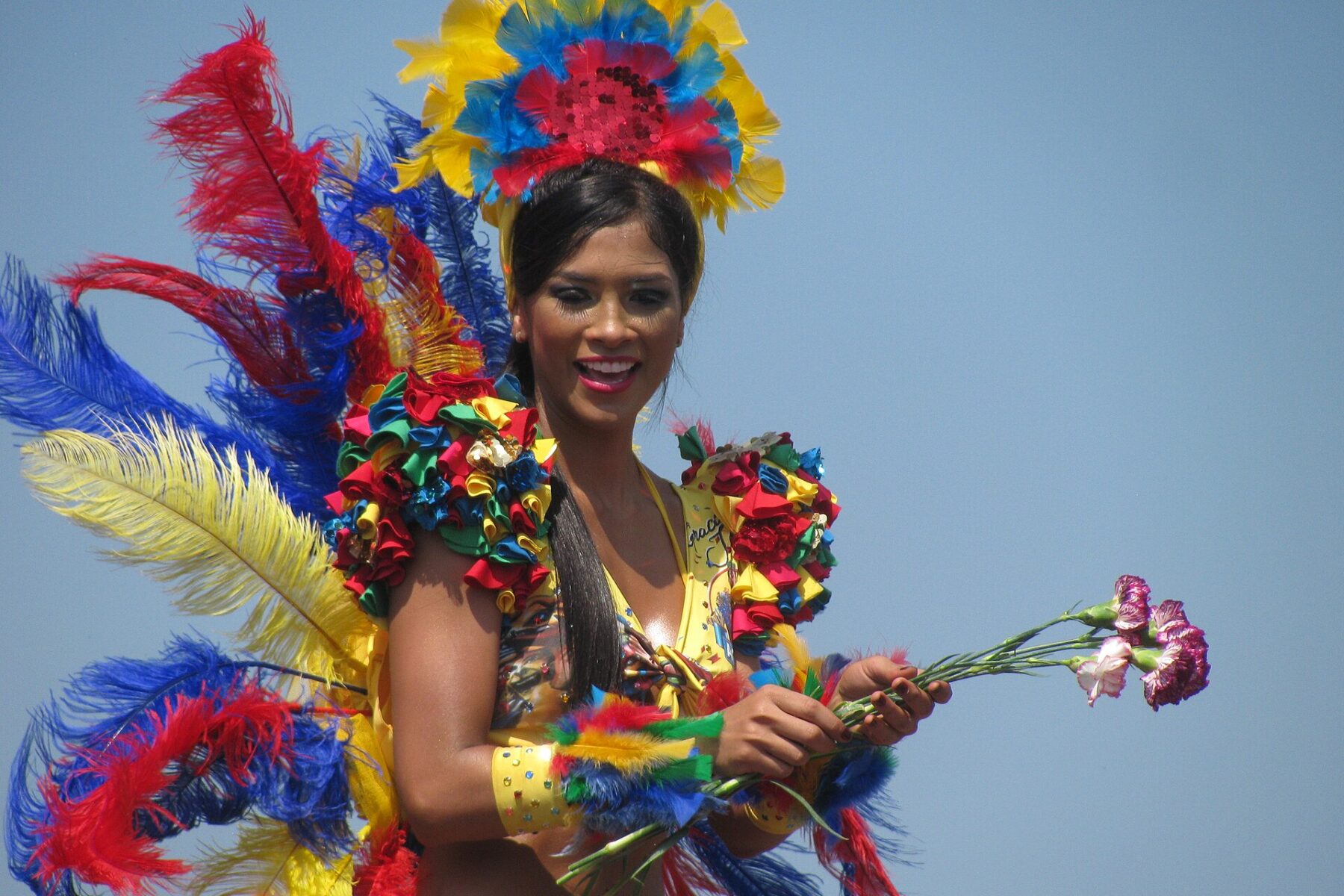 A smiling carnival dancer in a yellow costume with a red, blue and yellow feathered headdress holds carnations on a parade float