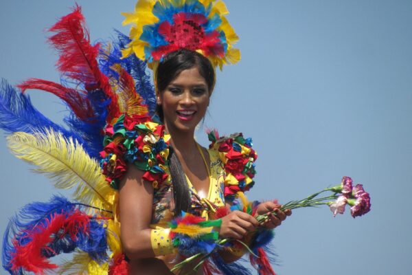 A smiling carnival dancer in a yellow costume with a red, blue and yellow feathered headdress holds carnations on a parade float