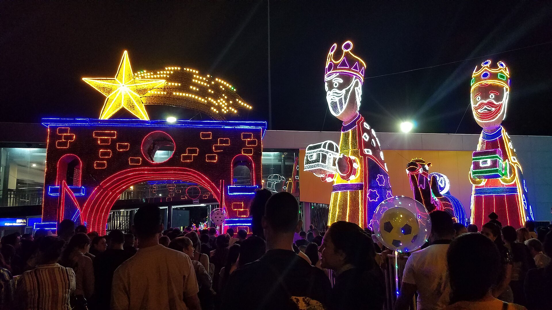 Illuminated light figures of the Three Kings, a camel, a star and a nativity arch glow at night above a crowd of visitors