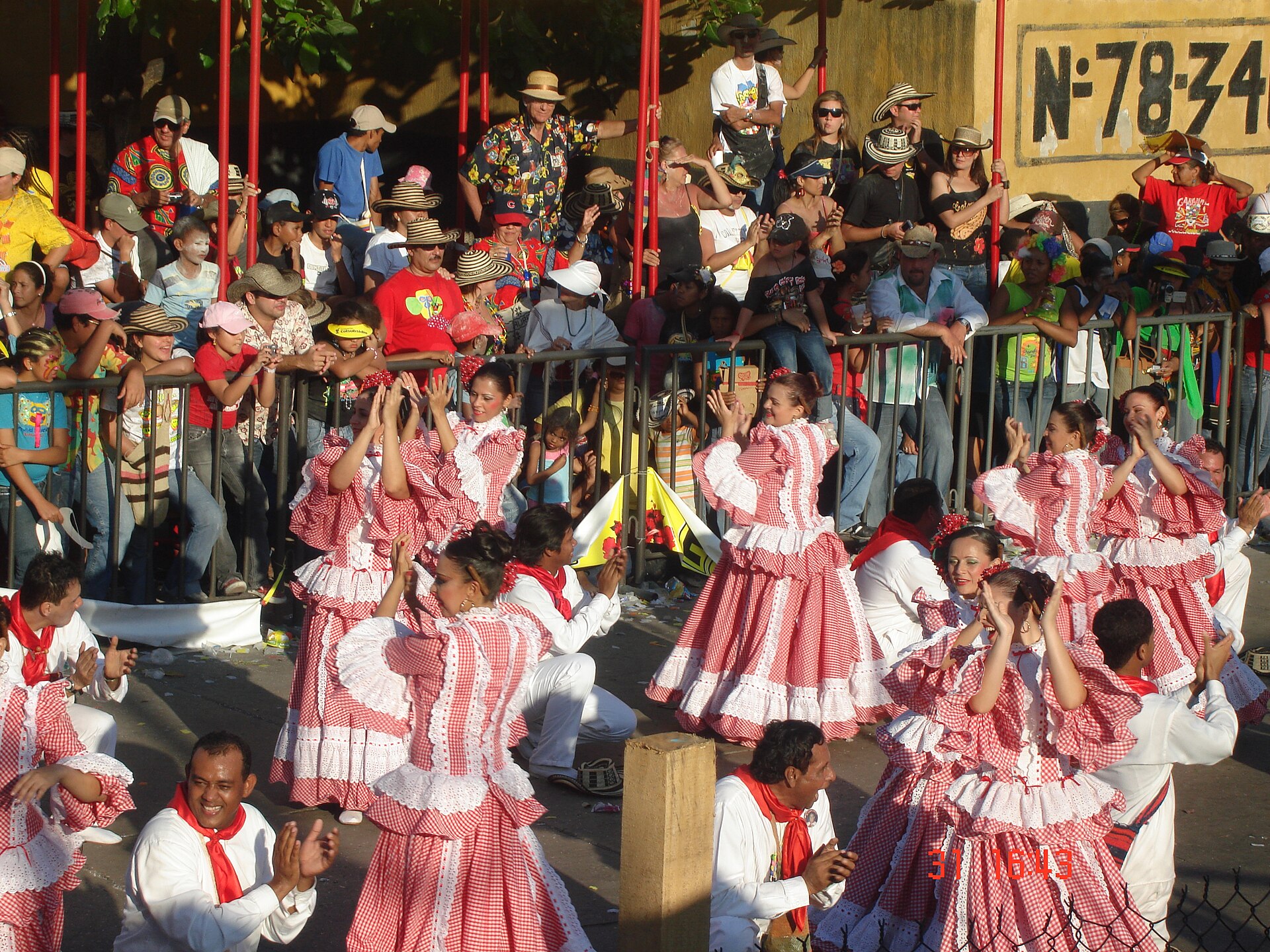 Women in red and white checked dresses and men in white with red neckerchiefs dance cumbia in a street as spectators in hats watch behind railings