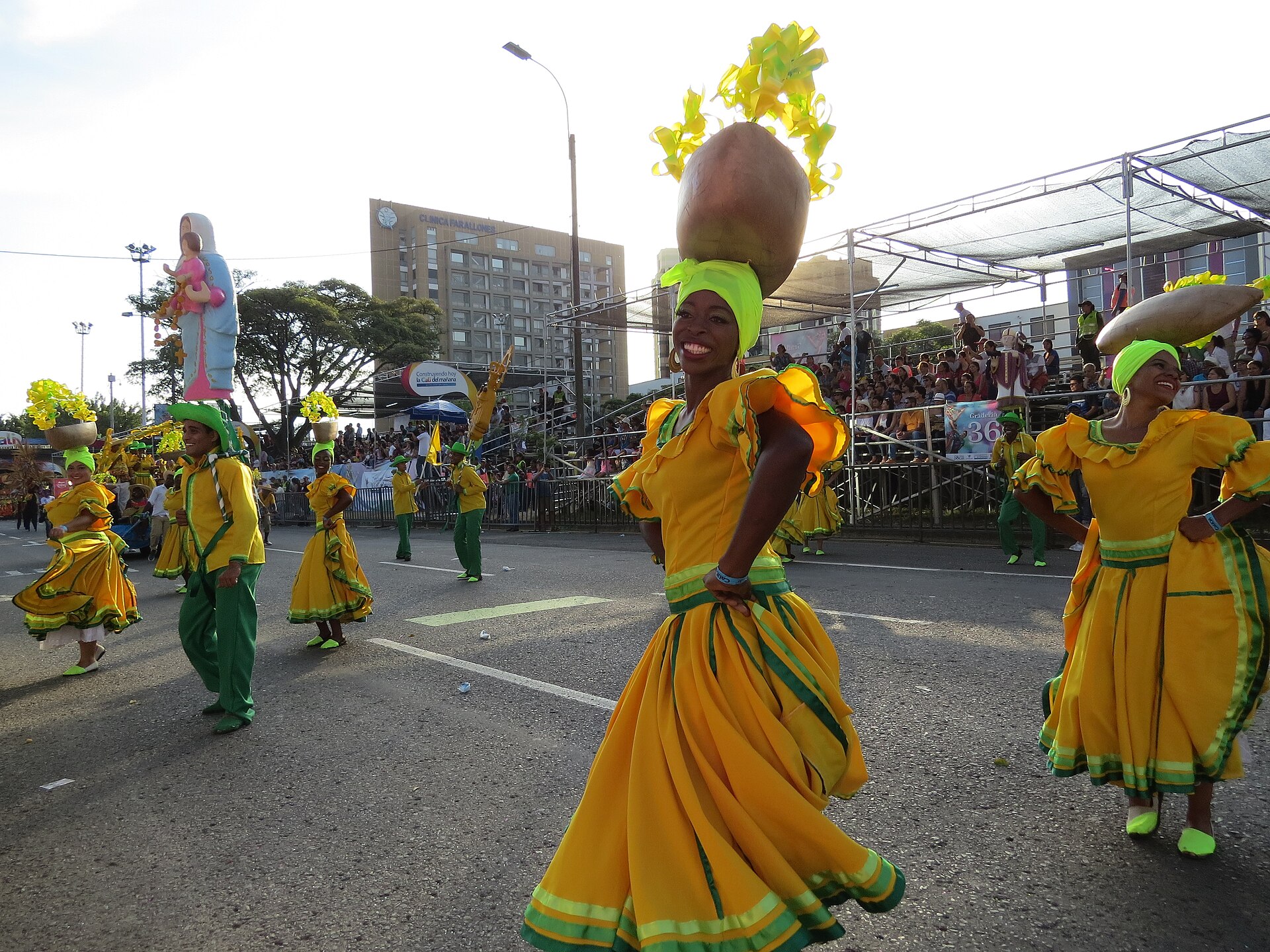 Smiling dancers in yellow and green dresses carry gourds on their heads along a street lined with grandstands