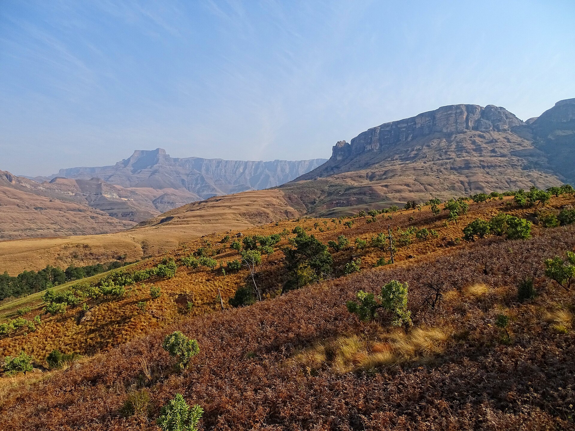Grassy slopes with scattered bushes rise towards rocky cliffs and the long rock wall of the Amphitheatre under a hazy blue sky