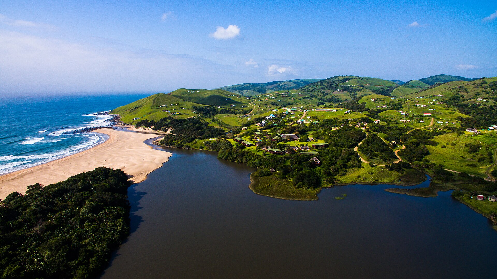 Aerial view of a river mouth and sandy beach on the Indian Ocean, with green hills dotted with houses behind a lagoon