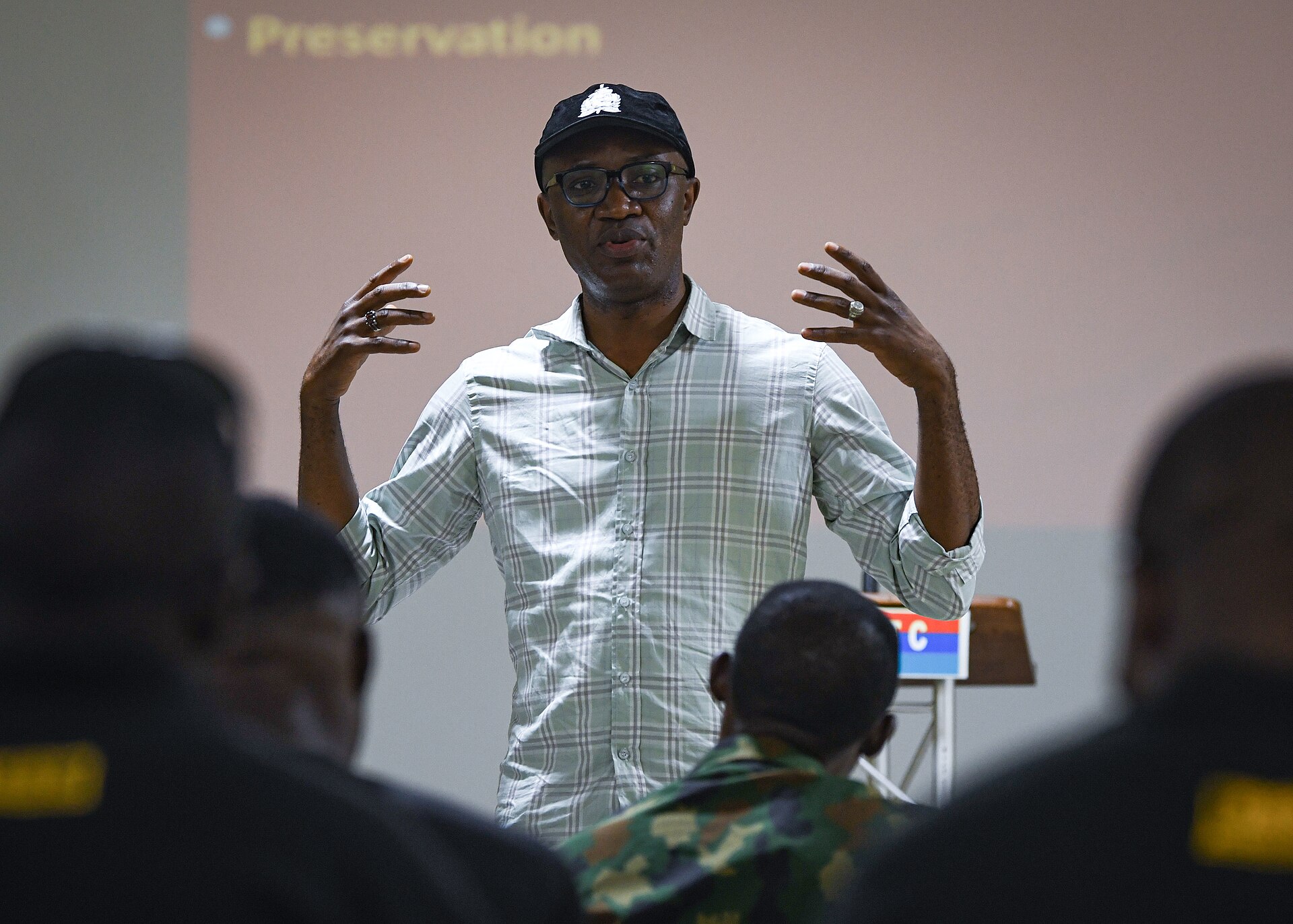 A man in a cap and checked shirt gestures while speaking to seated trainees in police and military uniforms in a classroom