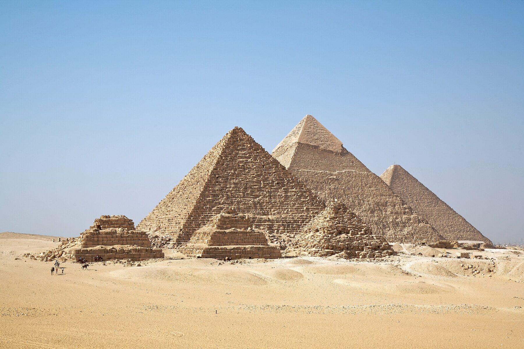The three Giza pyramids and three smaller queens' pyramids rise from golden desert sand under a clear blue sky, with a few camels and riders in the foreground