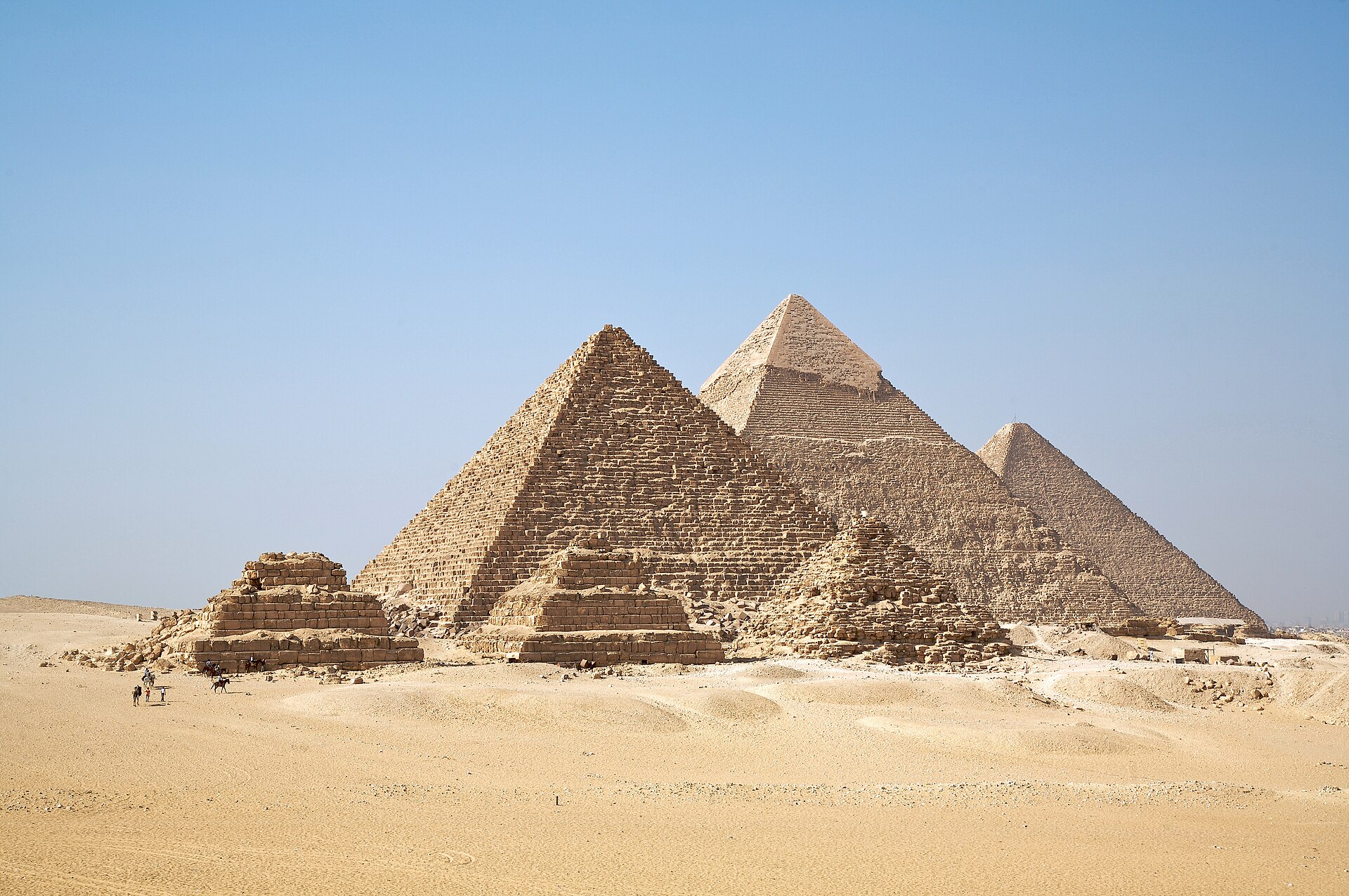 The three Giza pyramids and three smaller queens' pyramids rise from golden desert sand under a clear blue sky, with a few camels and riders in the foreground