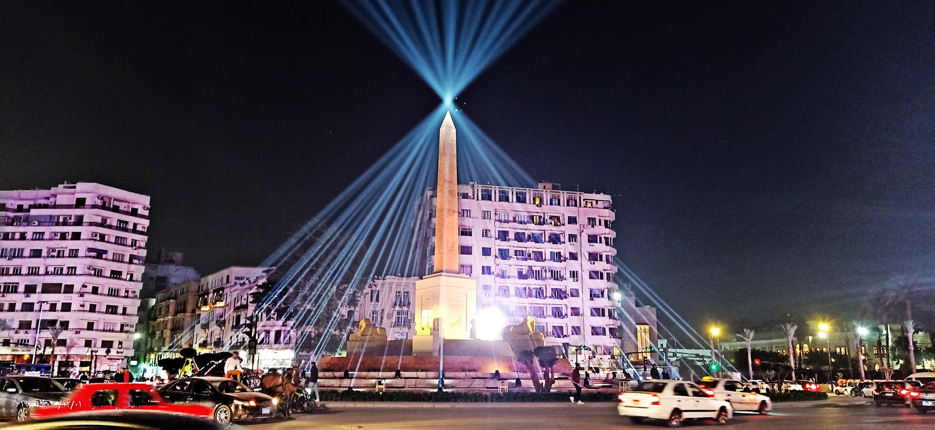 Tahrir Square at night with its central obelisk lit by blue searchlight beams, apartment blocks behind and cars circling the square