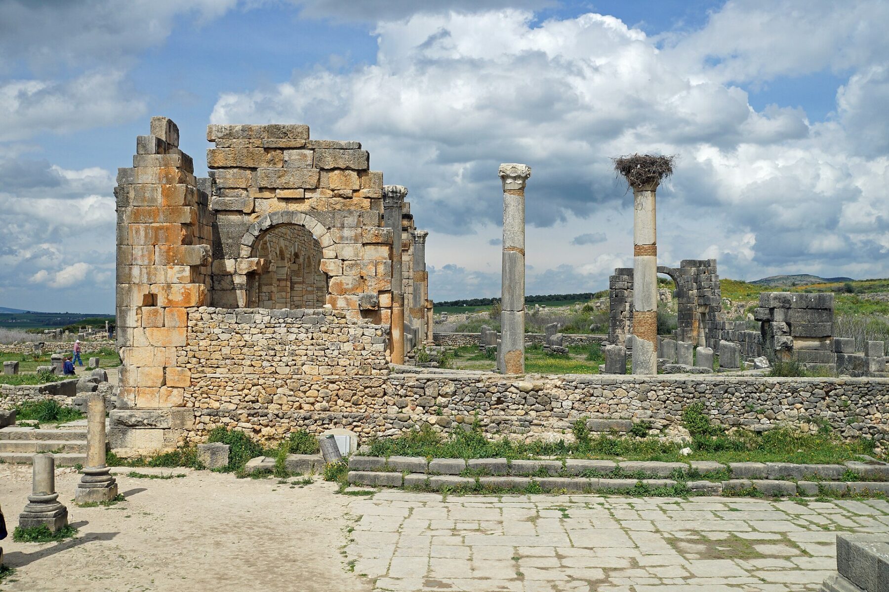 Ruined stone walls with an arch and tall Roman columns, one topped by a stork nest, stand on a paved square under a cloudy blue sky