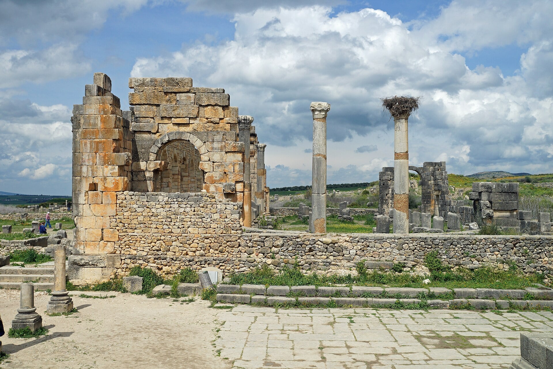Ruined stone walls with an arch and tall Roman columns, one topped by a stork nest, stand on a paved square under a cloudy blue sky